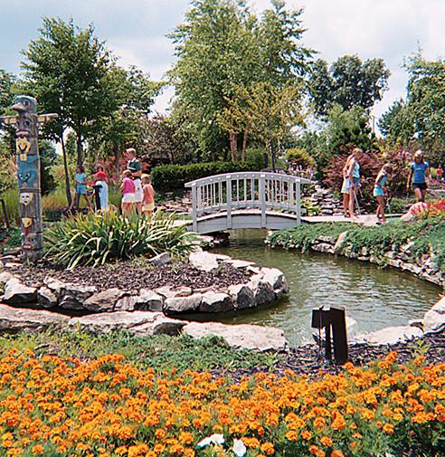 PowerPlay miniature golf course with adults are in background, blooming flowers in front, and bridge in center of shot.